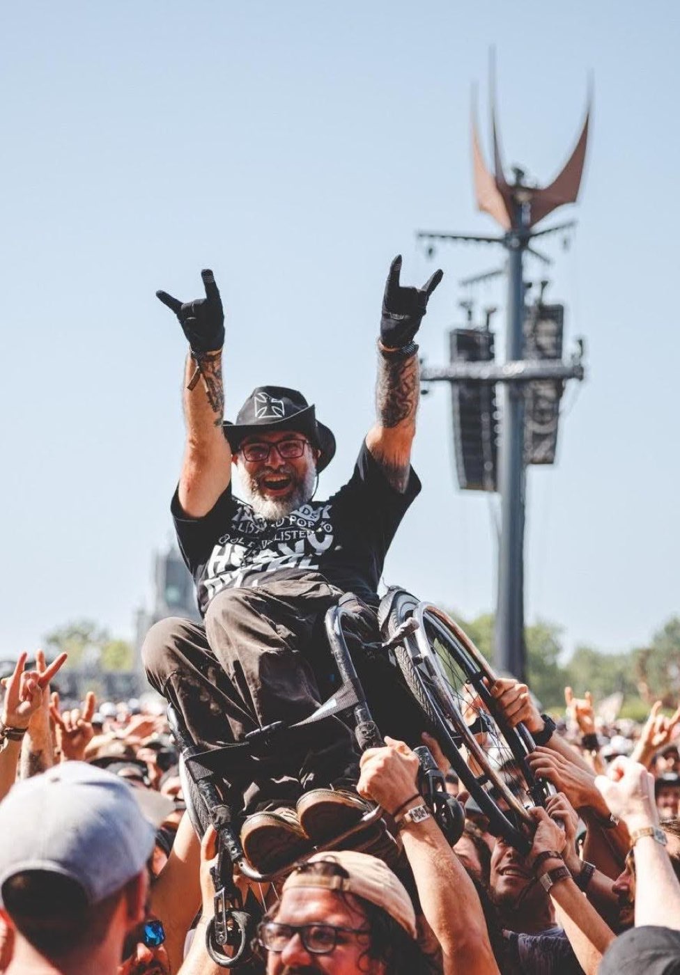 Slam en fauteuil roulant, les cornes levées, devant le totem du Hellfest